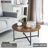 An industrial-style round coffee table from BONGO, featuring a rustic dark wood-grain top and a black metal base with an X-stretcher. The table is shown in a living room setting in front of a gray sofa, styled with books, a vase, and a coffee cup.
